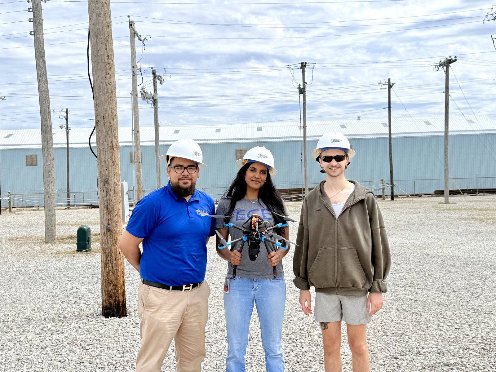 Derek Marin Martinez, Gwenith Madhan and Wyatt Fulton in hard hats holding the drone with the monitoring module mounted, at a utility yard